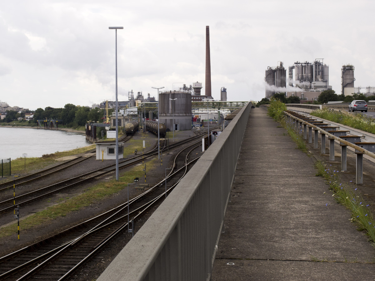 Fußweg zwischen Leitplanke und Brückengeländer über ein Industriegebiet am Rheinufer.
